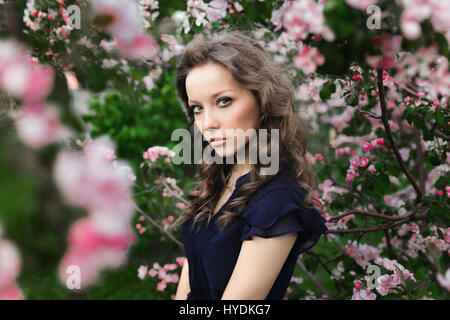 Clouse-up portrait of a young curly-haired girl in a blue dress standing among a blossoming apple tree Stock Photo