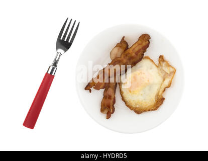 Top view of a fried egg with two strips of bacon on a plate with a fork to the side isolated on a white background. Stock Photo