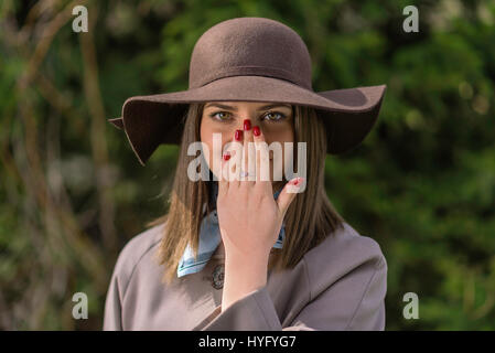 Young woman in hat and raincoat is posing in the park during a sunny spring day. She covered her mouth and nose with her hand with red nails. Stock Photo