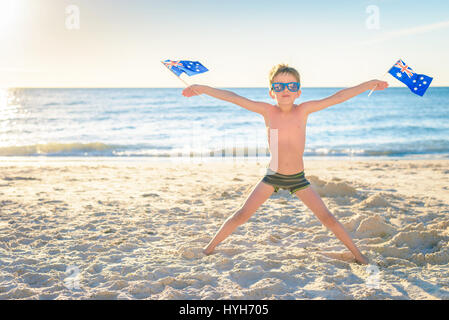 Cute active smiling boy standing on the beach and holding Australian flags on Australia day Stock Photo