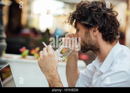 handsome elegant man sitting outside at a restaurant table, working and using laptop while smoking cigarettes Stock Photo