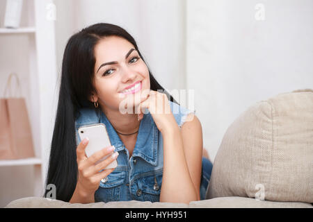 Positive young woman lying on the couch and uses a mobile phone Stock Photo