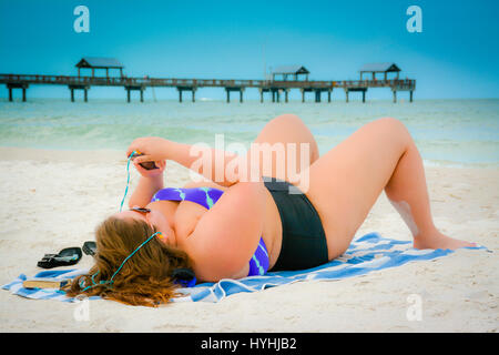 Plus size woman in two piece swimsuit lying on Clearwater Beach, FL holding Smart phone with earphones and Pier 60 in the Gulf of Mexico Stock Photo