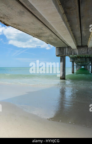 View underneath Pier 60 at Clearwater Beach, with concrete pilings and blue-green waters with white sandy beach on the Gulf of Mexico in FL Stock Photo