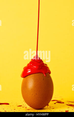 red paint falling on a brown egg placed on a yellow background splashed by the same paint Stock Photo