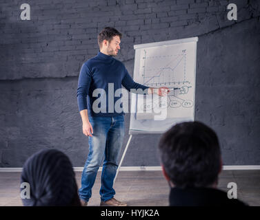 Young speaker man talking to audience during conference. He is giving a lecture and showing graph on flipchart.  Business presentation concept Stock Photo