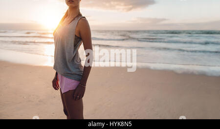 Cropped shot of fitness woman standing on the beach. Sporty female on the sea shore in morning for workout. Stock Photo