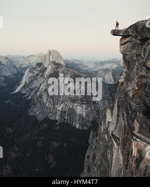 A fearless hiker is standing on an overhanging rock enjoying the view towards famous Half Dome at Glacier Point in Yosemite National Park, California