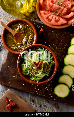 high-angle shot of some earthenware bowls with zucchini spaghetti, italian pesto sauce and chopped tomato, and a cruet with olive oil on a rustic wood Stock Photo