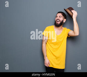 Portrait of a happy young man with beard laughing and holding hat on gray background Stock Photo