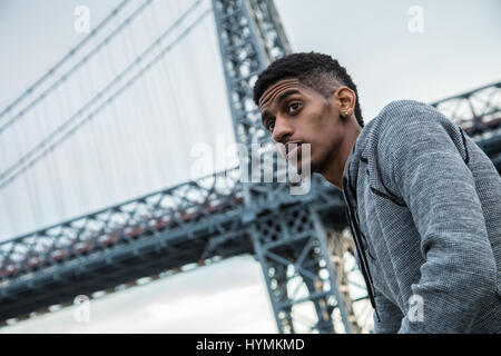 A portrait of a young, black man, pensively standing along New York City's East River and Williamsburg Bridge. Stock Photo