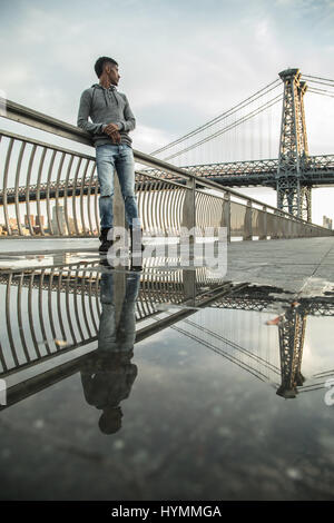 A young, black man walks along New York City's East River with Brooklyn's Williamsburg Bridge in the background. Shot during the Autumn of 2016. Stock Photo
