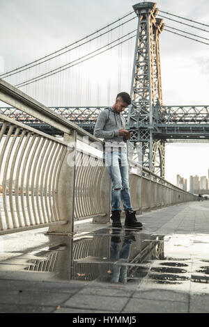 A young man texts infront of New York City's Williamsburg Bridge at sunset. Shot in Brooklyn, New York City during the Autumn of 2016. Stock Photo