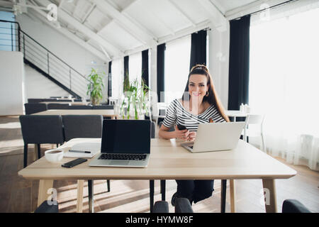 Smiling Attractive Woman Working on Laptop in Cafe, while holding a phone. Stock Photo
