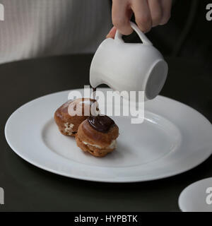 A jug with hot chocolate and two eclairs with cream on a plate on a table in a cafe. girl is pouring the eclair by chocolate Stock Photo