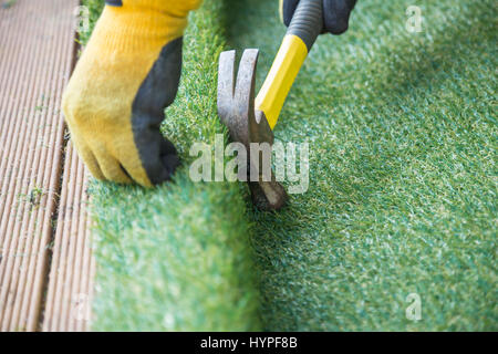 Artificial grass, turf installation alongside decking. A hammer in being used to nail the turf into place. A yellow work glove can be seen. Stock Photo