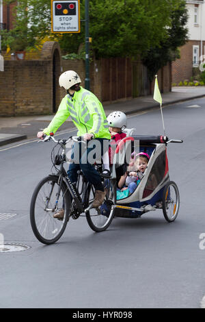 Woman cyclist on bike / bicycle with + 3 children; Co Pilot child seat with helmet & towing cycle Chariot trailer with two / 2 kids with helmets. UK. Stock Photo