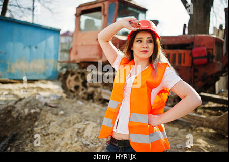 Engineer builder woman in uniform waistcoat and orange protective helmet against bulldozer. Stock Photo