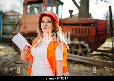 Engineer builder woman in uniform waistcoat and orange protective helmet against bulldozer. Stock Photo