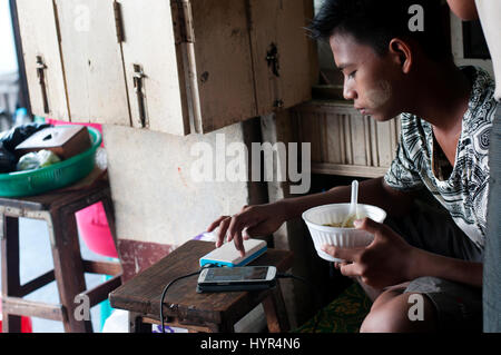 Myanmar (Burma). Yangon. A young man working in the market checks his phone whilst eating his lunch. Stock Photo