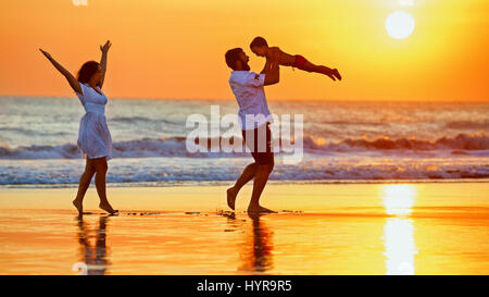 Happy family - father, mother, baby son walk with fun along edge of sunset sea surf on black sand beach. Active parents and people outdoor activity on