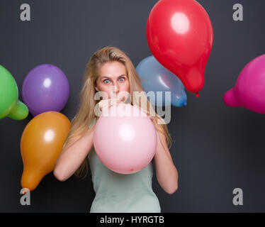 Portrait of a beautiful blond woman blowing up balloon on gray background Stock Photo