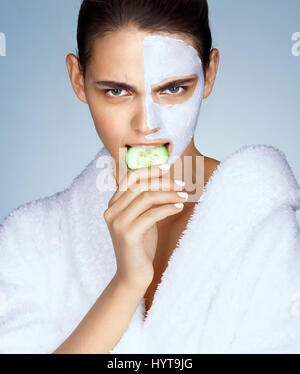 Young girl with rejuvenating mask bites a piece of cucumber. Photo of brunette in white bathrobe on blue background. Wellness and Spa concept Stock Photo