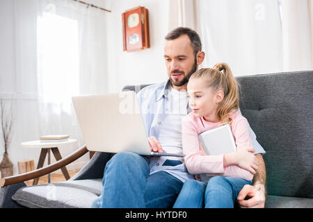 Father and daughter sitting embracing on sofa and using laptop Stock Photo