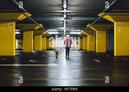 Girl wearing white hat and red jacket walking in the public garage with small yellow dog Stock Photo