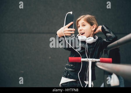 Girl wearing black leather jacket and white headphones standing on the scooter taking selfie with smart phone outdoor Stock Photo