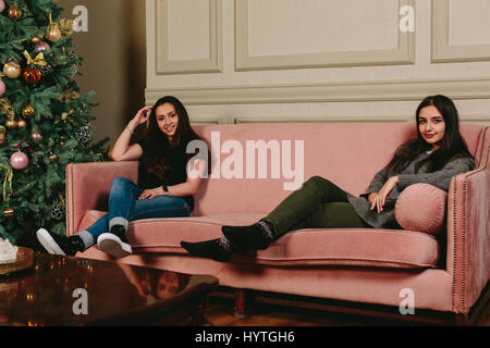 Two beautiful young girls on a sofa near the Christmas tree. Studio horizontal portrait. Stock Photo