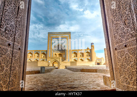 View to the arched old madrassa though the wooden door with oriental patterns. Khiva old town, Uzbekistan Stock Photo