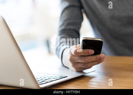 Close-up of incognito businessman in grey sweater sitting at wooden table next to laptop and holding black cell phone in one hand. Stock Photo