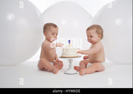 One year old twin brothers wearing diapers and eating their birthday cake. Shot in the studio with large, white balloons in the background. Stock Photo