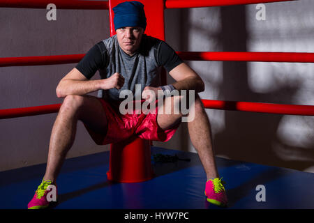 Athletic male boxer sitting near red corner of a regular boxing ring surrounded by ropes in a gym Stock Photo