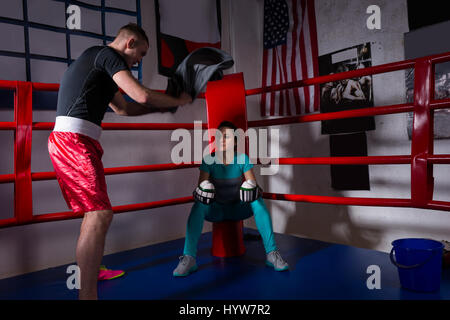 Young sporty female boxer in boxing gloves after battle sitting next coach near red corner of a regular boxing ring in a gym Stock Photo