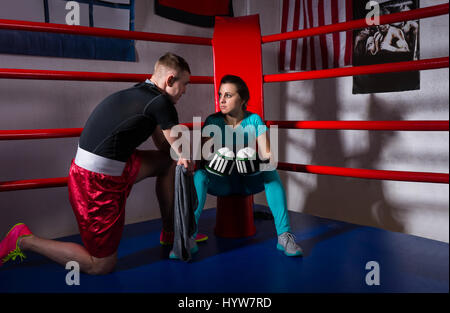 Young female boxer in boxing gloves after battle sitting next coach near red corner of a regular boxing ring in a gym Stock Photo