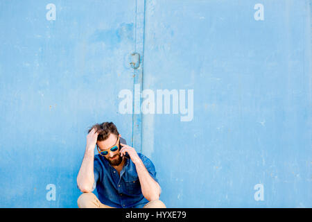 Man dressed casual in blue shirt sitting with phone on the blue wall background. Wide shot with copy space Stock Photo