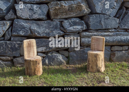Two handmade wood stools on the garden lawn with different natural stones wall as background Stock Photo
