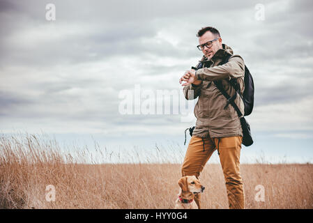 Hiker with dog standing in grassland and looking at watch Stock Photo