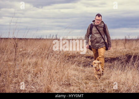 Hiker and small yellow dog walking in grassland on a cloudy day Stock Photo