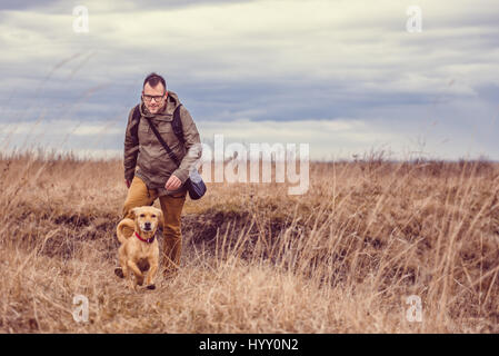 Hiker and small yellow dog walking in grassland on a cloudy day Stock Photo