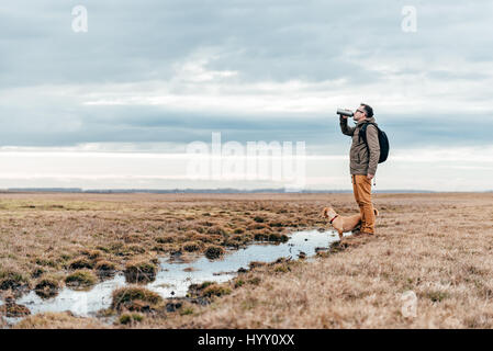 Hiker and dog standing by the pond in grassland on a cloudy day and drinking water Stock Photo