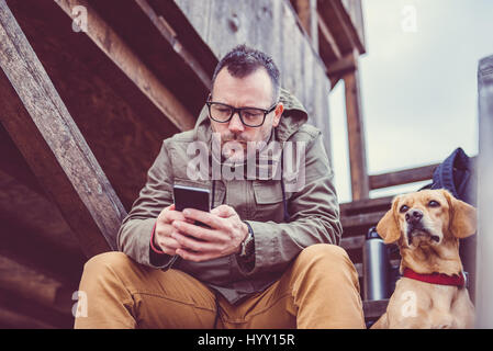 Hiker sitting on the stairs of hikers rest cabin with a dog and using smart phone Stock Photo