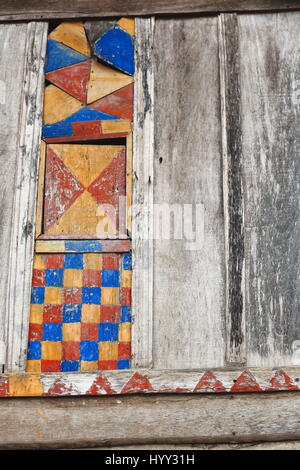 Colorfully yellow-red-blue painted wooden wall and paint-faded boards of a window with checkered decoration. Hamlet of the Akha Pala hill tribe-Pak Na Stock Photo