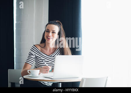 Portrait of woman laughing and looking away with a laptop in a cafe. Lady is sitting at the table near window with a cup. Stock Photo