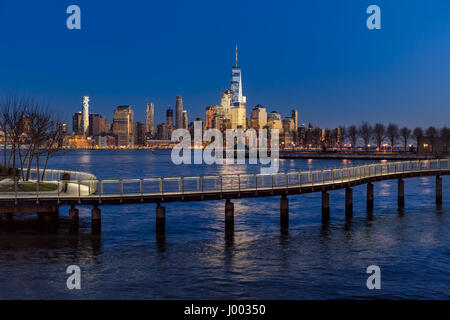 New York City Financial District skyscrapers at sunset and Hudson River from Hoboken promenade. Lower Manhattan skyline and pedestrian bridge Stock Photo