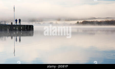 Two people stand on Waterhead Pier in a misty Windermere lake at Ambleside in England's Lake District National Park. Stock Photo