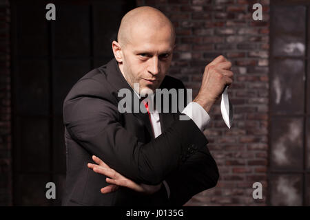 Silent killer, oriental martial arts in action. Bald contract murderer in suit and red tie holds combat knife. Fighting knife is a dangerous weapon in Stock Photo