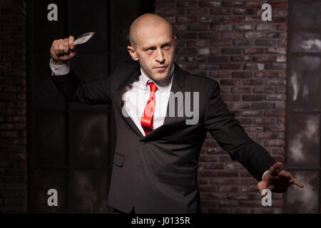 Silent assassin, oriental martial arts in action. Bald contract murderer in suit and red tie holds combat knife. Fighting knife is a dangerous weapon  Stock Photo